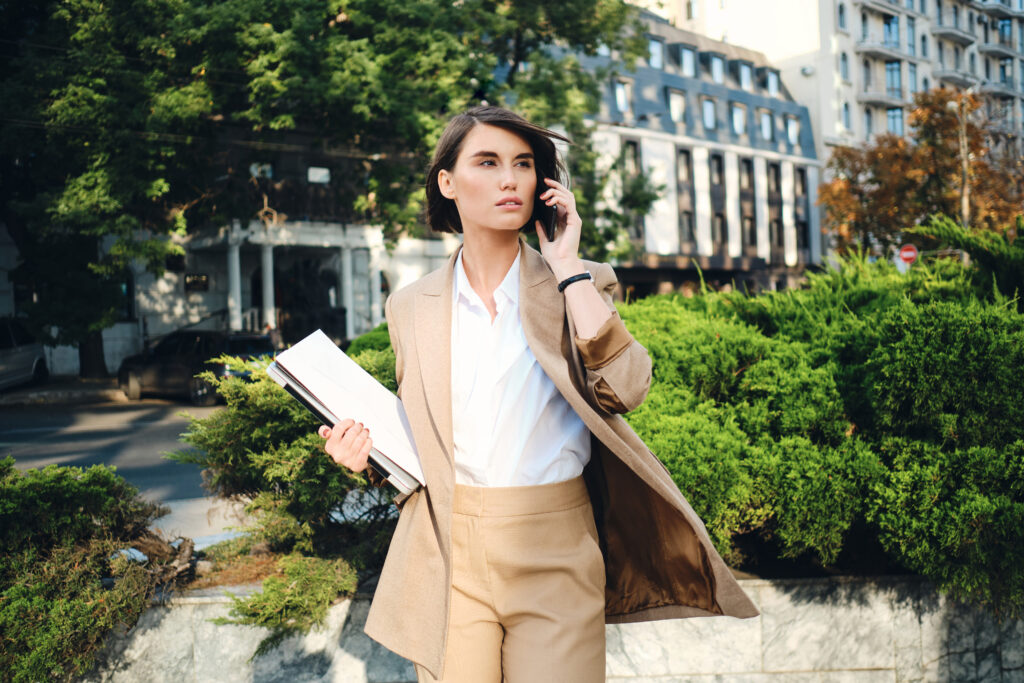 young attractive businesswoman beige suit with laptop thoughtfully talking cellphone city street