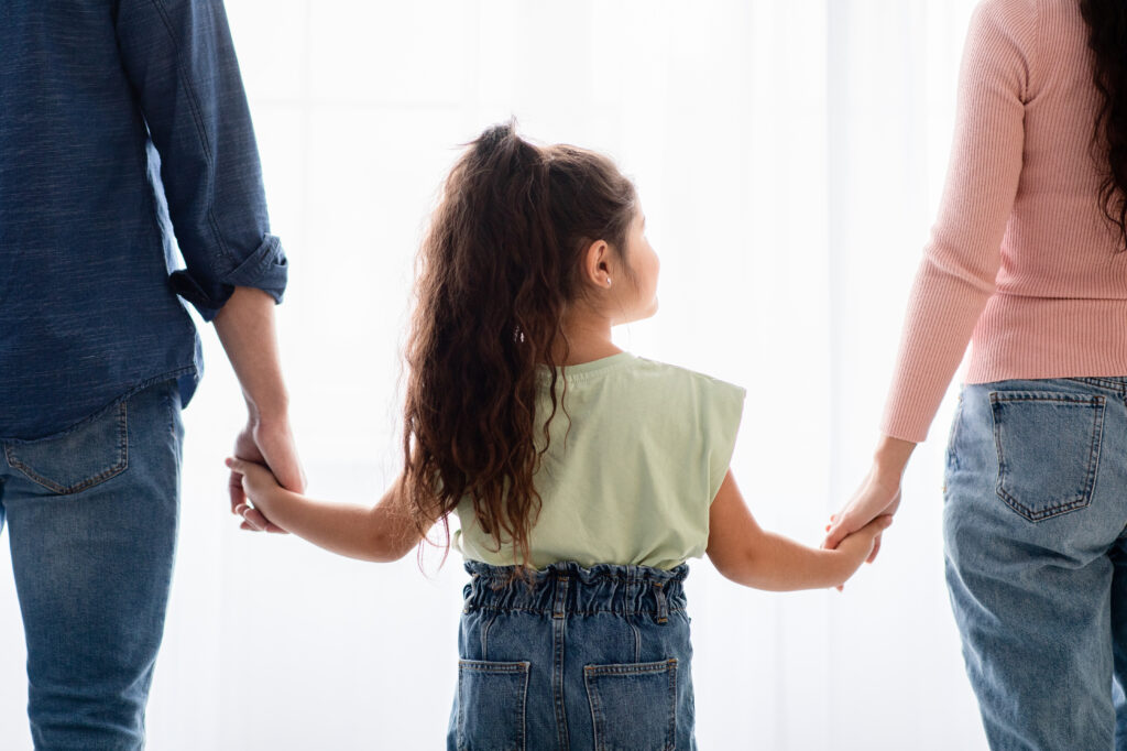 family support concept rear view little girl holding hands with mom dad standing near window home closeup shot parents female child bonding together cropped image